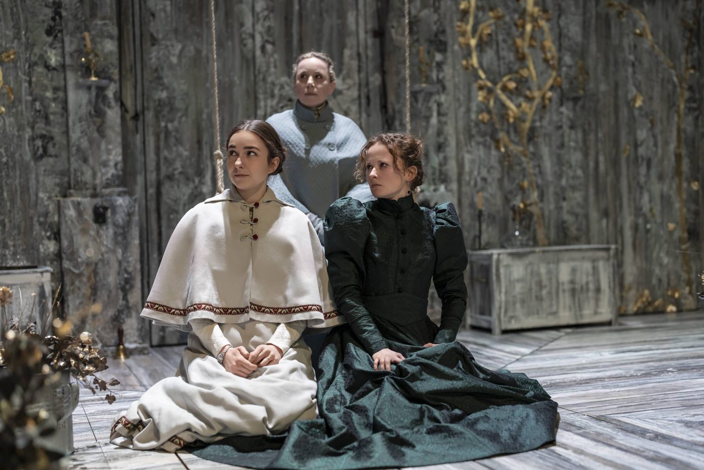 Ruby Thompson as Irina, Michelle Terry as Olga, and Shannon Tarbet as Masha in Three Sisters. They are sat on the floor of the stage, wearing typical Russian costumes from the period, with elegant dresses and capes to keep out the cold.