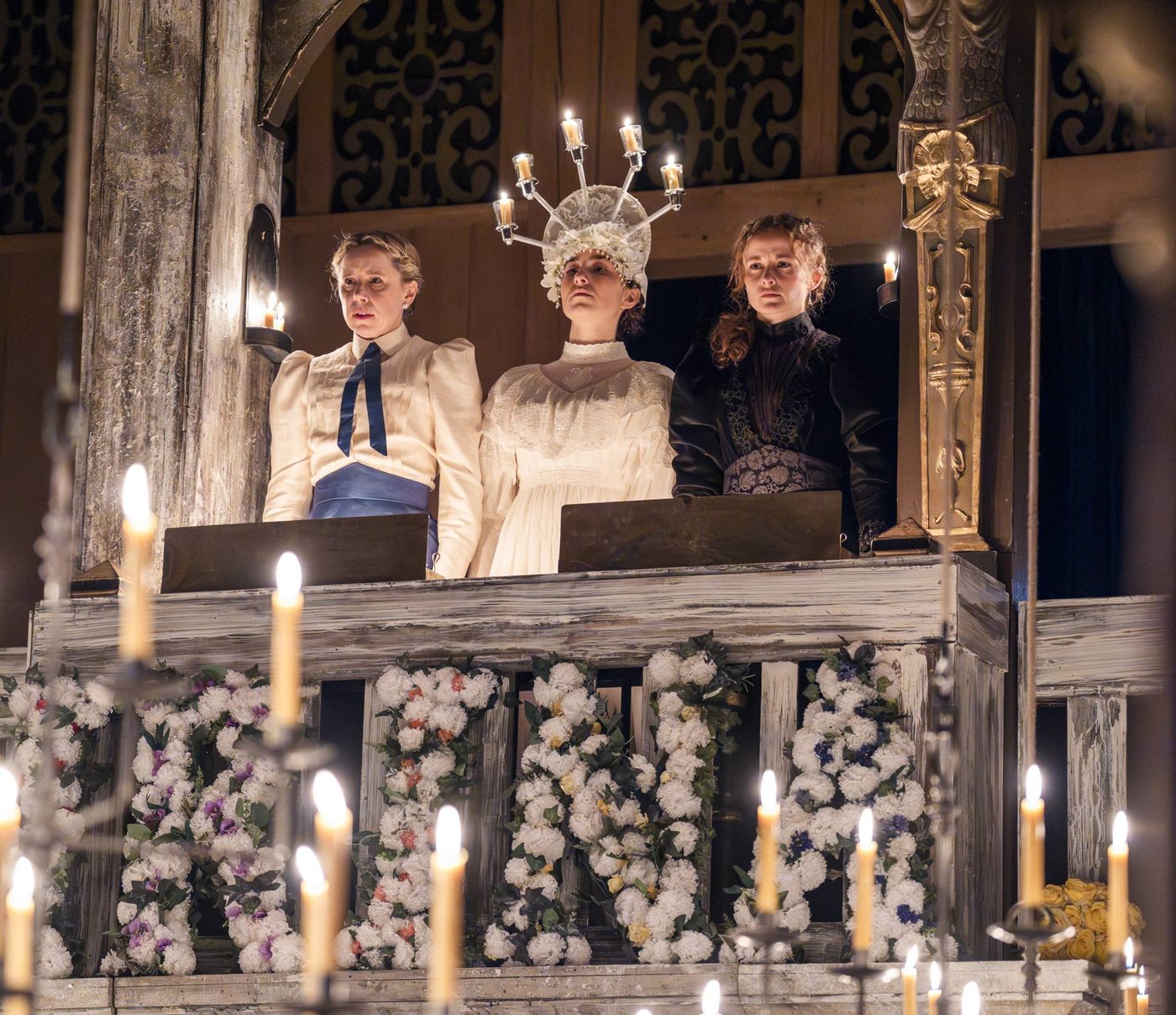 Michelle Terry as Olga, Shannon Tarbet as Masha and Ruby Thompson as Irina in Three Sisters. Olga wears a blouse and blue skirt, Irina wears a white lacey dress with a tiara made from lit candles, and Masha wears a dark black dress with her hair over one shoulder.