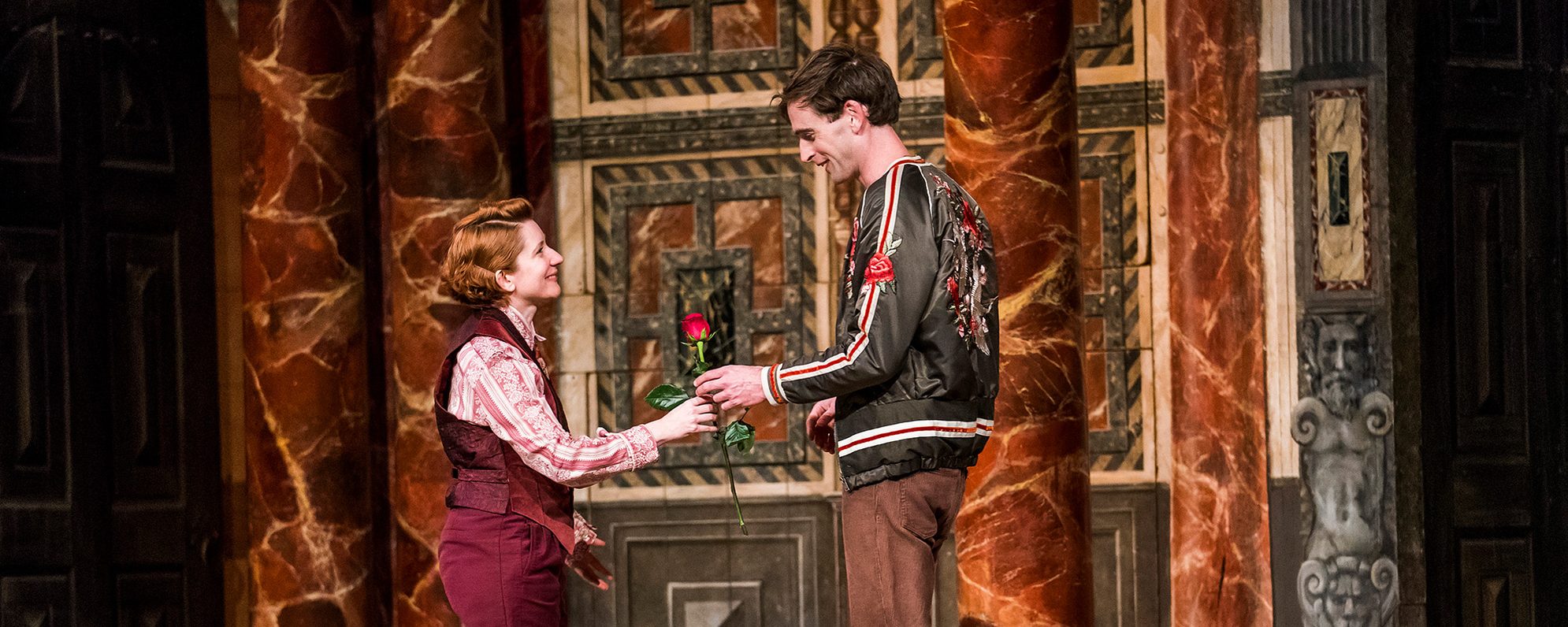 An actor hands another actor a red rose gift