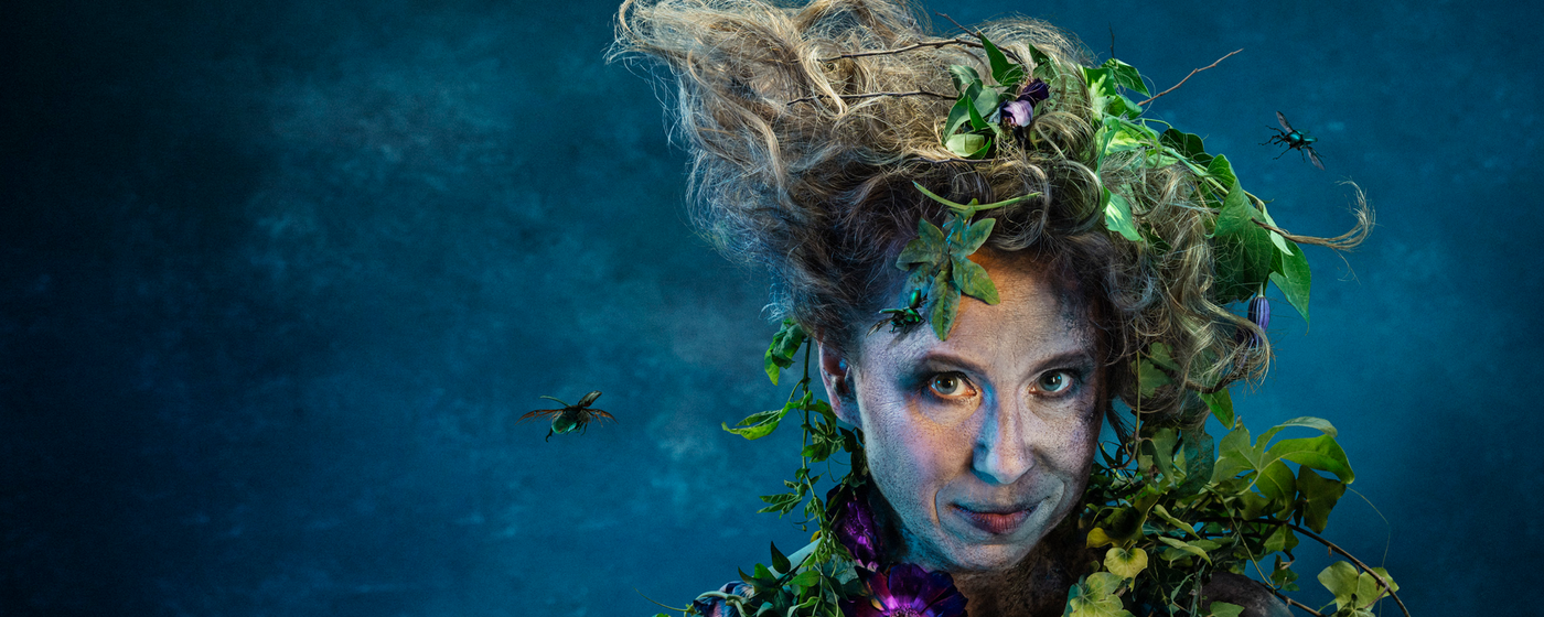 A close up of a smirking woman against a blue backdrop. She is covered in dirt and foliage and has insects flying around her unruly hair.